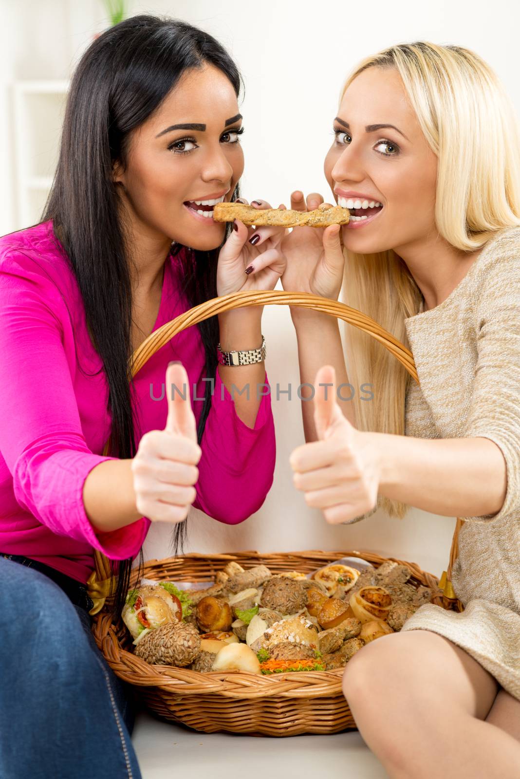 Brunette and blonde girl are sitting on the couch, raised their thumbs and looking at the camera with her mouth open about to bite a piece of pastry each with its end. Among them is woven basket with pastries.