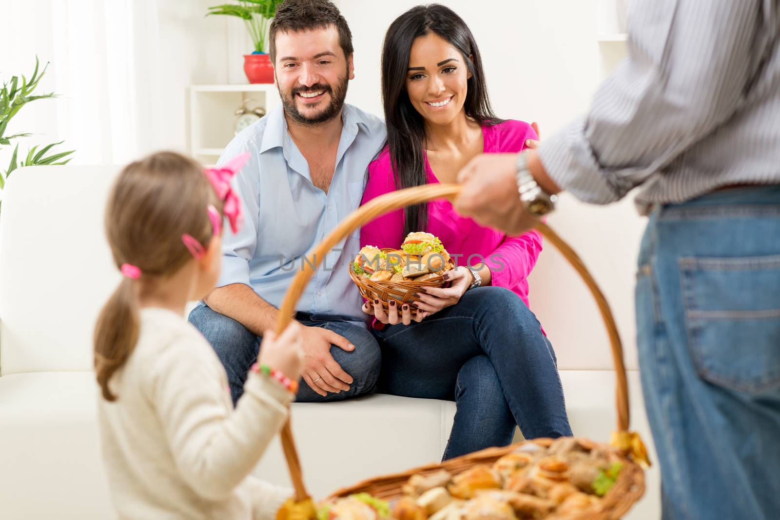 Young parents are sitting on the couch and looking at little daughter, who with her grandfather wearing wicker basket of pastries. A little girl and an older man were photographed from the back.