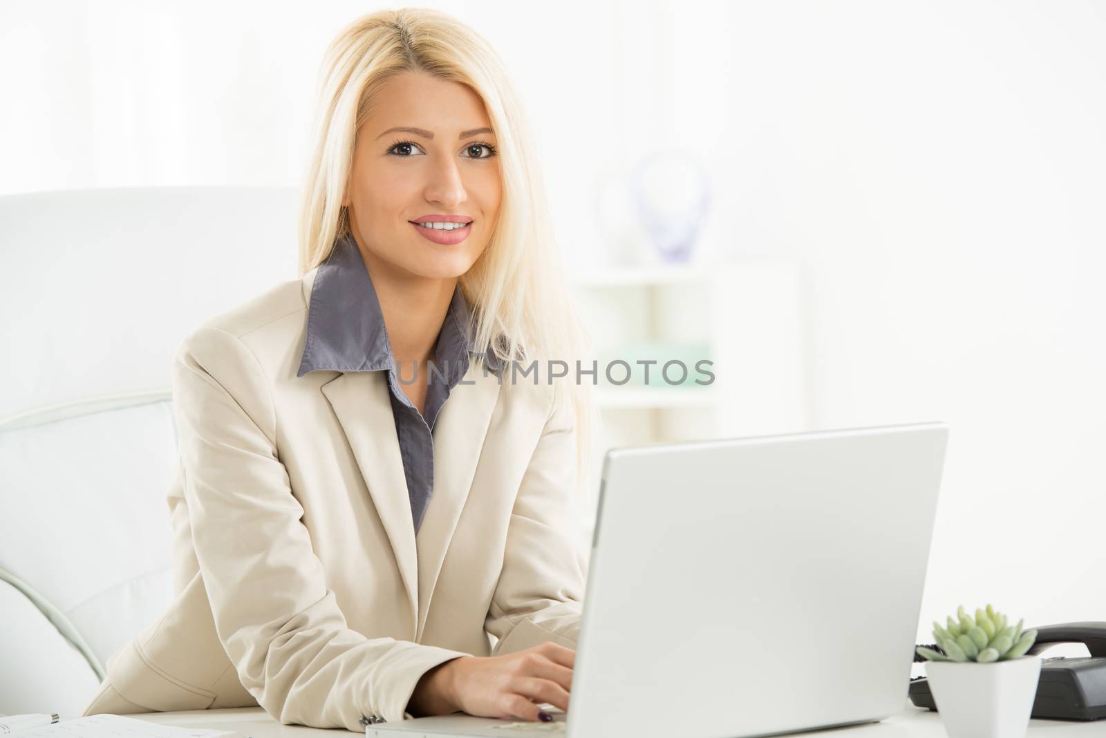 A young blonde businesswoman, elegantly dressed sitting at a desk in front of laptop in her office with a smile on her face looking at the camera.