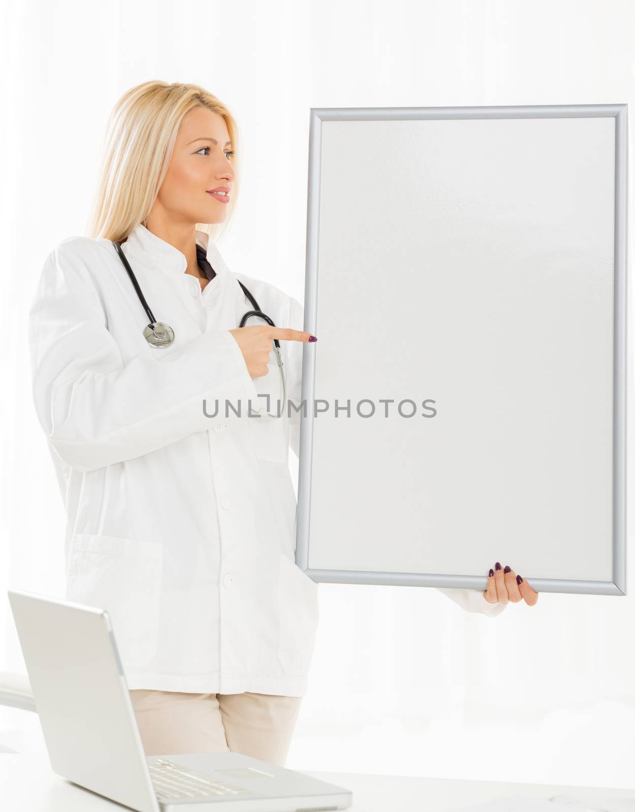 A pretty blonde girl in a white coat with a stethoscope over neck, standing in front of a table with a laptop, holding a blank notice board at which points a finger.