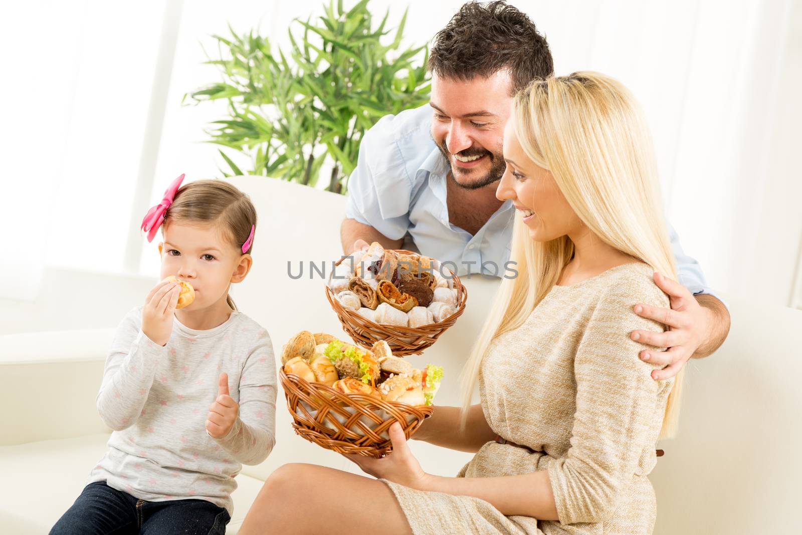 Young parents hold in their hands woven basket with delicious salty and sweet pastries. Beside them sit on the couch their little daughter eating pastries and holding thumb-up.