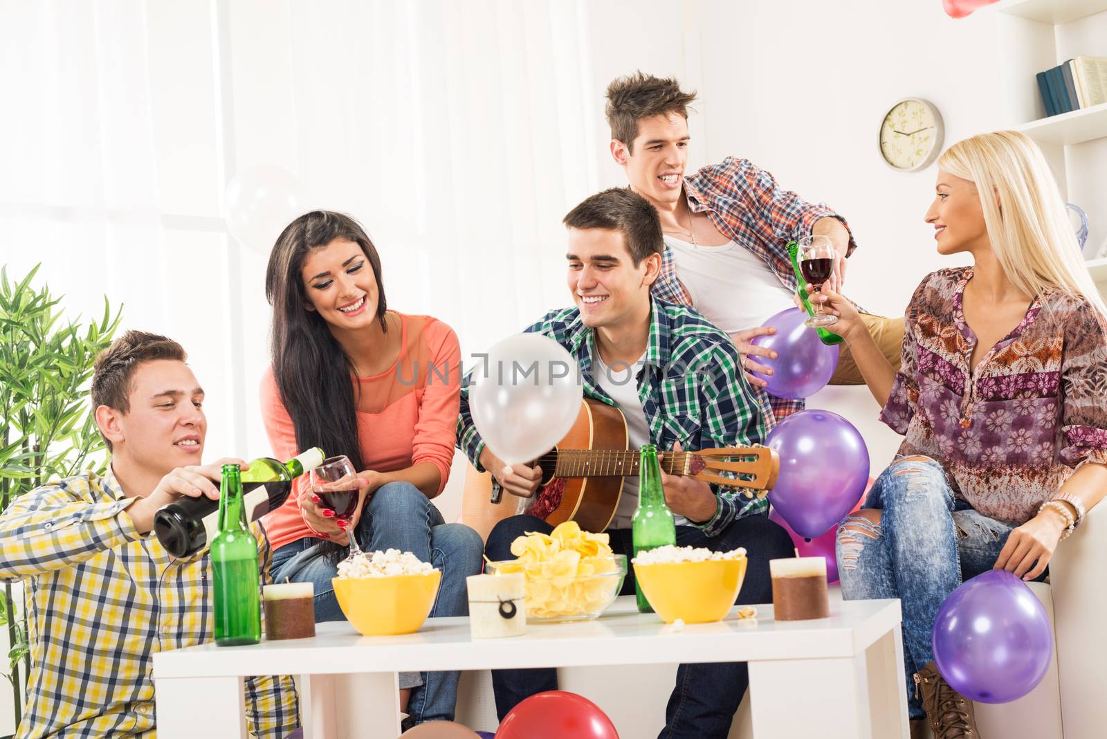 A small group of young people at the home party, enjoy the sounds of the acoustic guitar, drinking wine and beer.