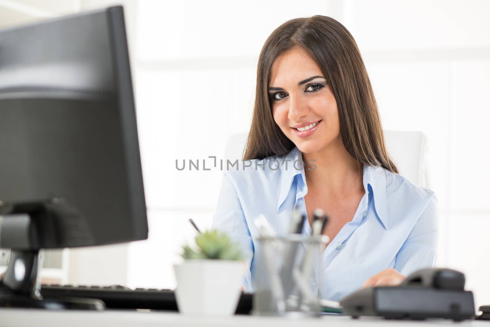 Pretty young business woman sitting at an office desk in front of the monitor, with a smile on her face looking at the camera.