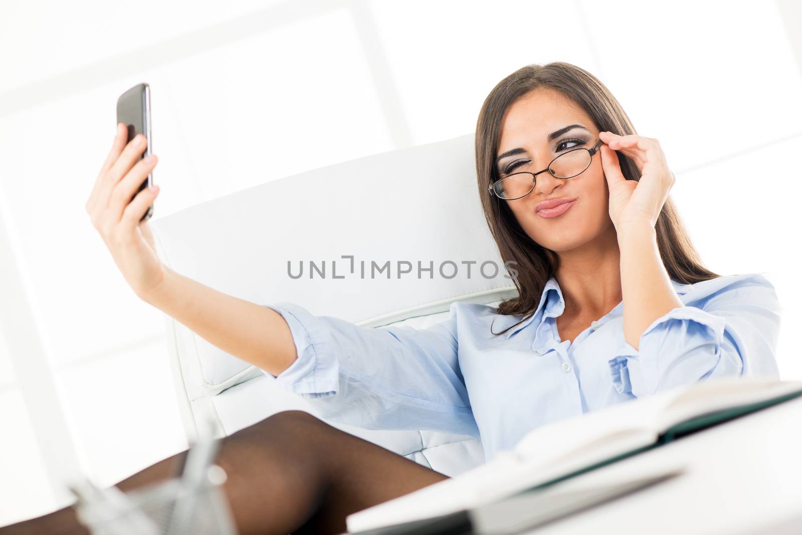Pretty young businesswoman with glasses, sitting in the office with her feet up on the desk, photographing herself with a cell phone.