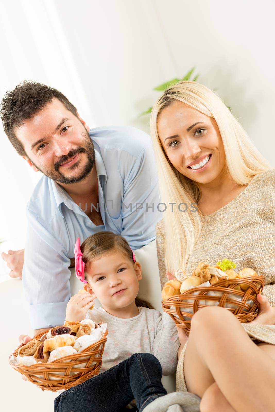 Young parents hold in their hands woven basket with delicious salty and sweet pastries. Beside them sit on the couch their little daughter eating pastries.