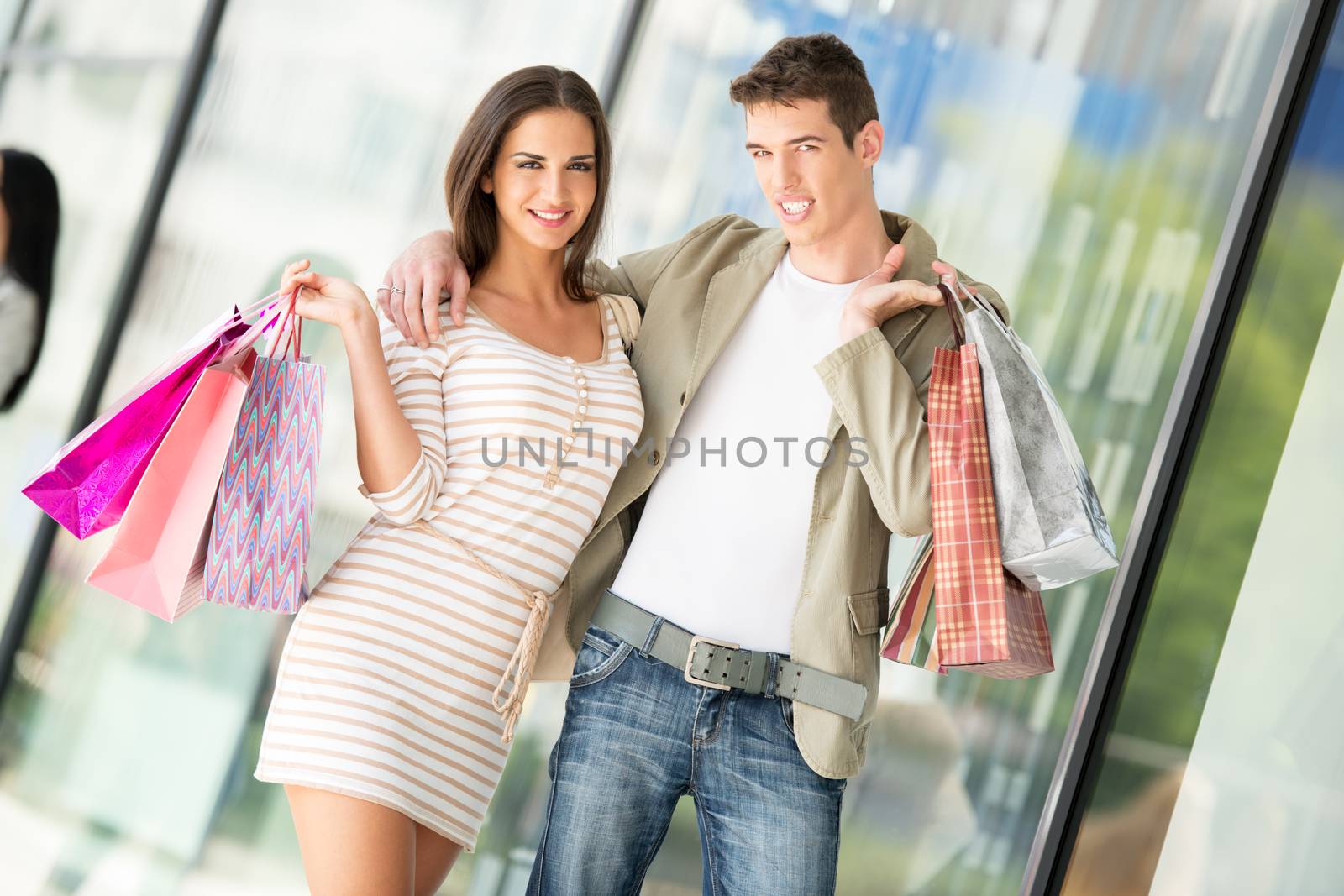 Young happy couple in shopping passes in front of window shopping mall carrying bags in their hands.