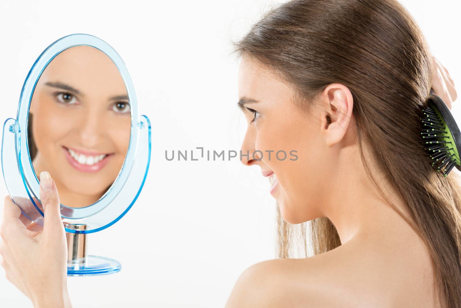 Girl holding a mirror in which looks and combing her long brown hair. Reflection of girl in the mirror looking at the camera with a smile.
