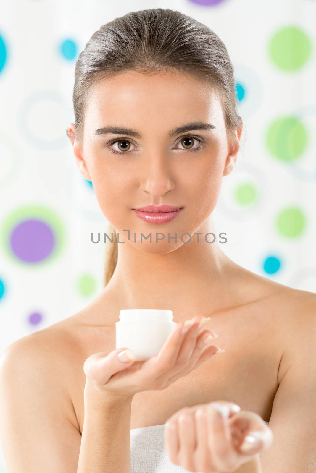 Beautiful young girl with a towel wrapped around her chest, holding cream in her hand and with a slight smile looking at the camera.