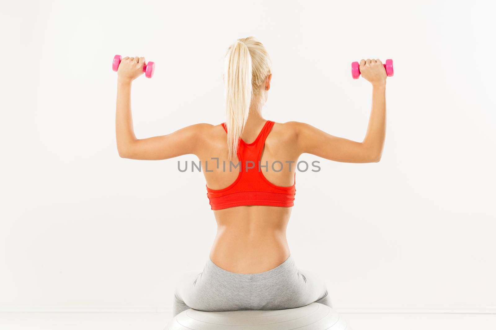 Cute sporty Young girl sitting to pilates ball and exercising with dumbbells. Rear view.