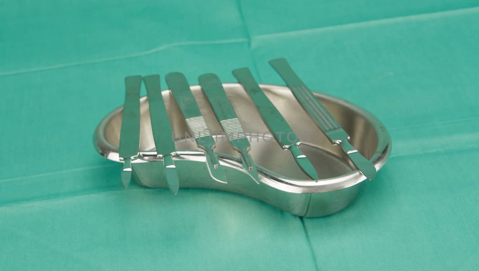 Many types of scalpel in kidney shape bowl on green fabric.