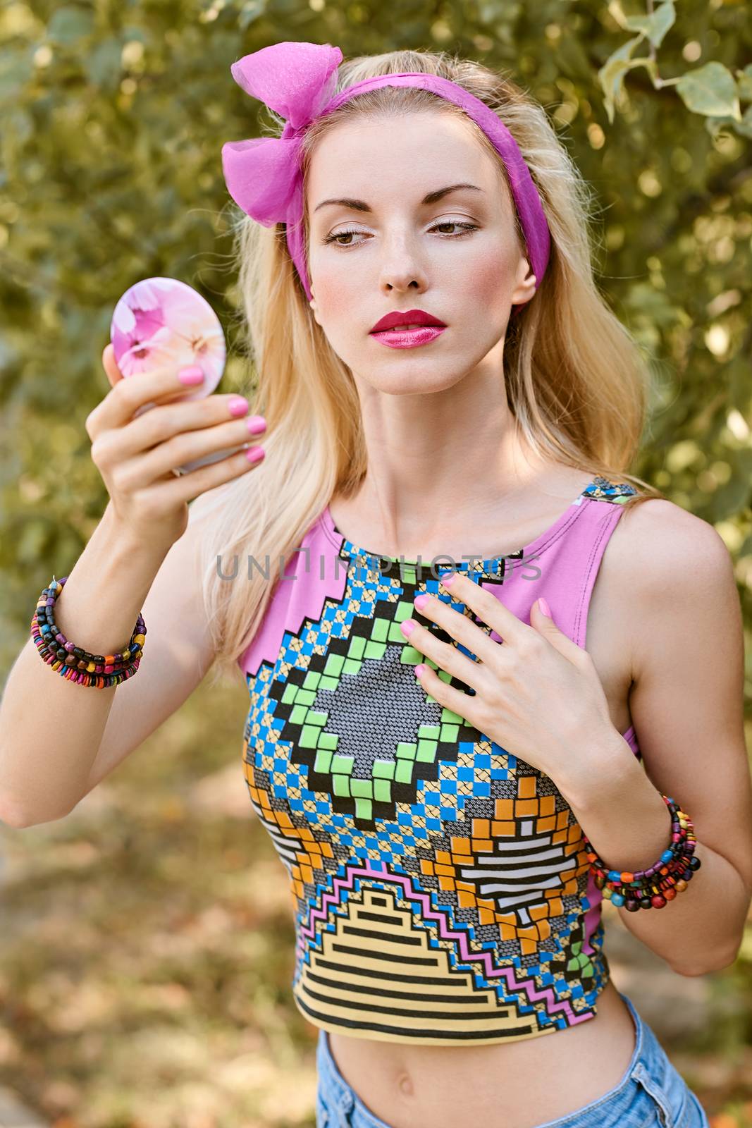 Beauty portrait stylish playful woman primping with mirror in park, people, outdoors. Attractive hipster happy pretty blonde girl with bow, fashionable top. Relax in summer garden, lifestyle, bokeh