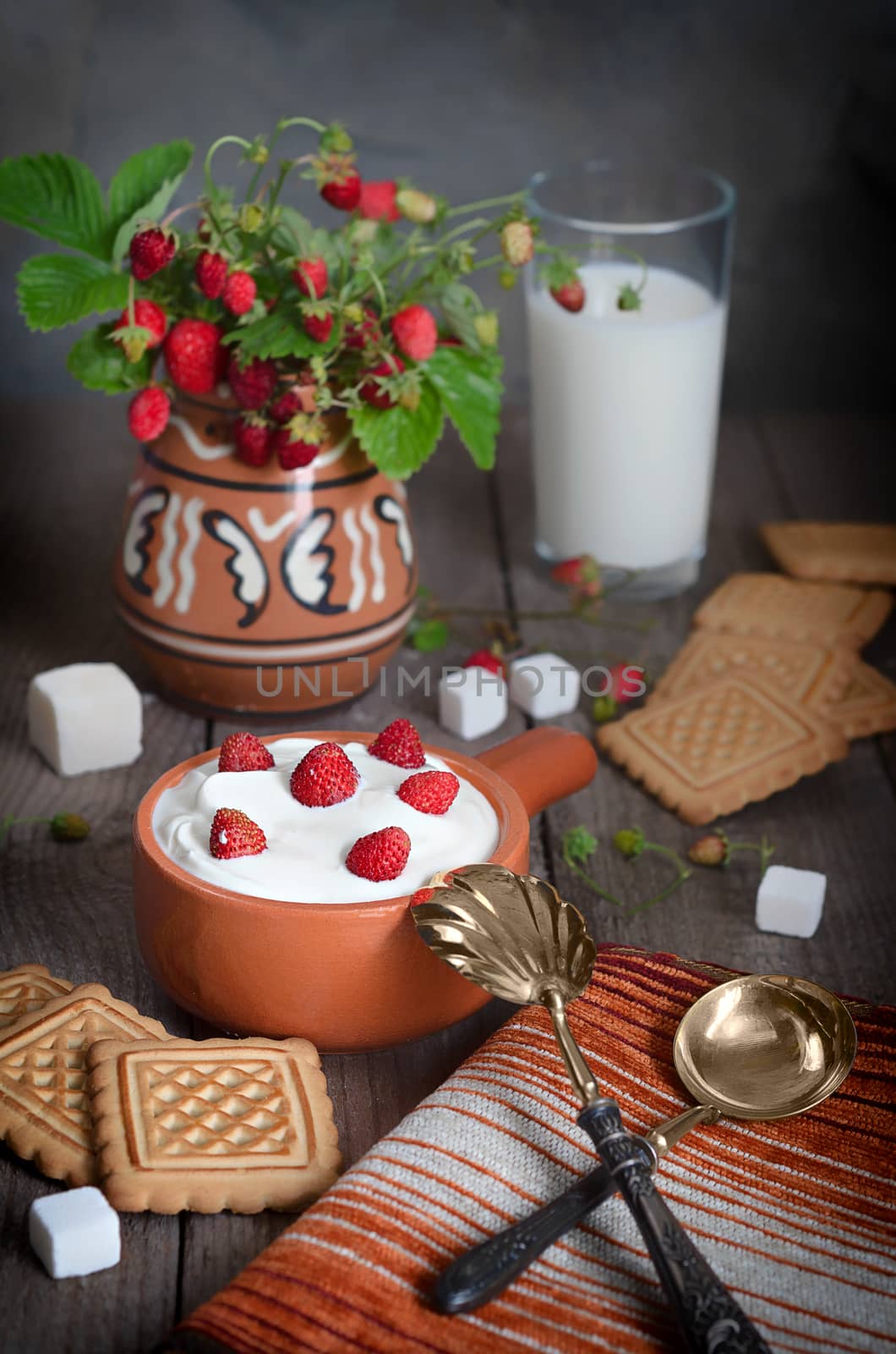 Strawberry with cream in a ceramic Cup, cookies and milk in glass on old wooden surface. Bouquet with strawberries in a ceramic vase and antique spoons on a napkin. The rustic style.