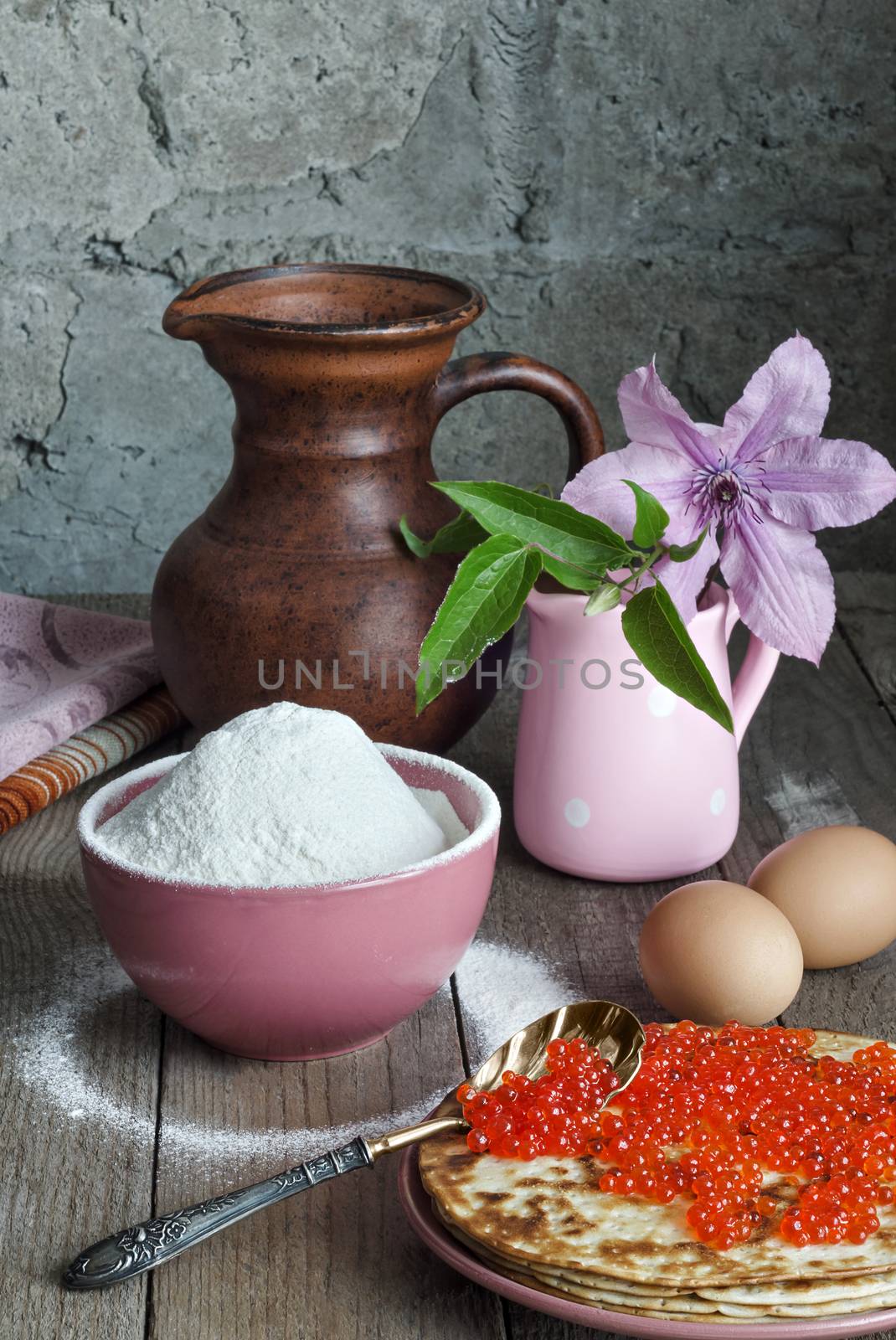 Pancakes with red caviar on a plate. Antique caviar spoon. In a bowl, flour, and eggs on a grey background and rough wooden surface. A pitcher and a vase with flowers in the background.
