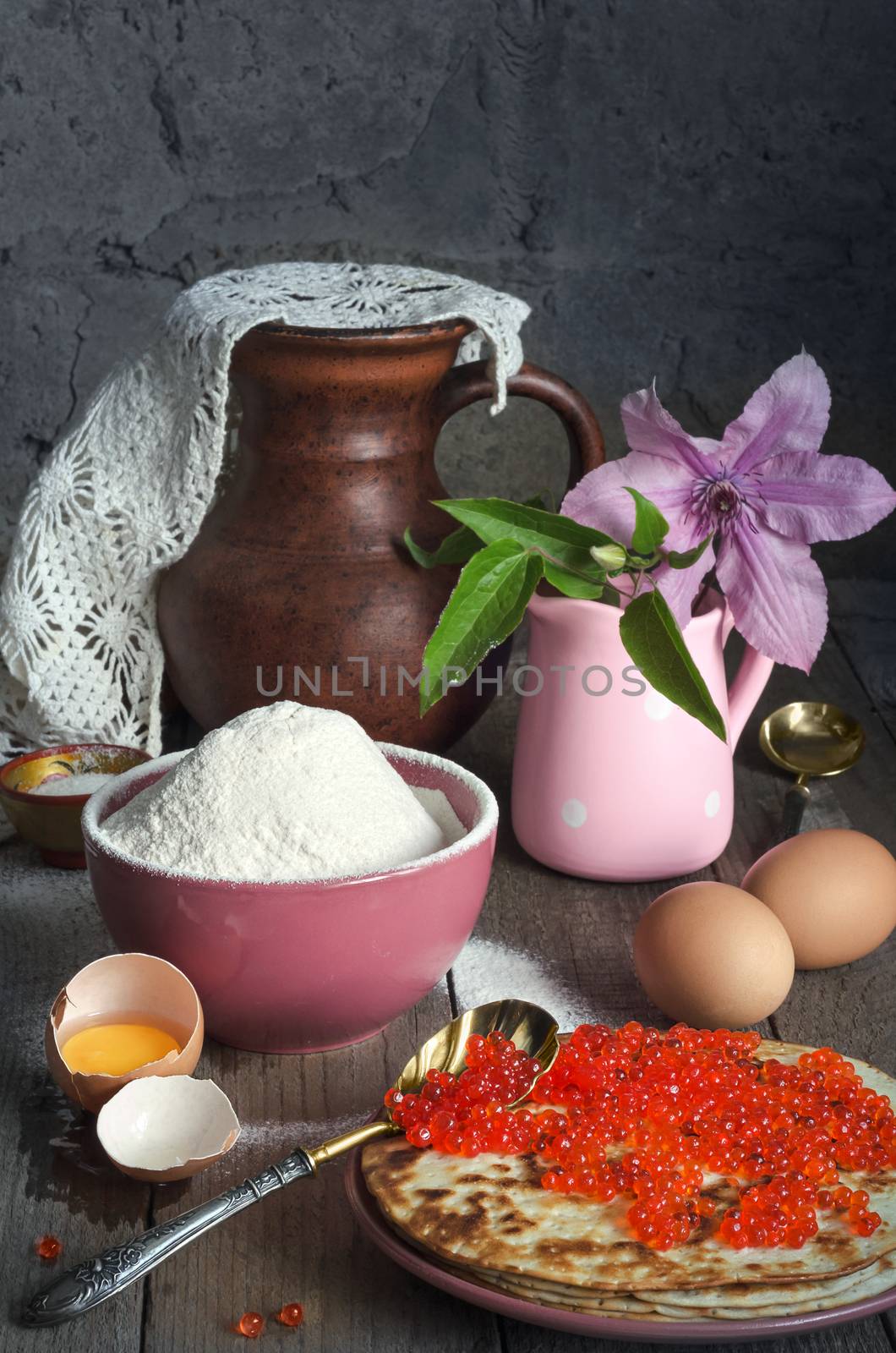 Pancakes with red caviar on a plate. Antique caviar spoon. In a bowl, flour, and eggs on a grey background and rough wooden surface. A pitcher and a vase with flowers in the background.
