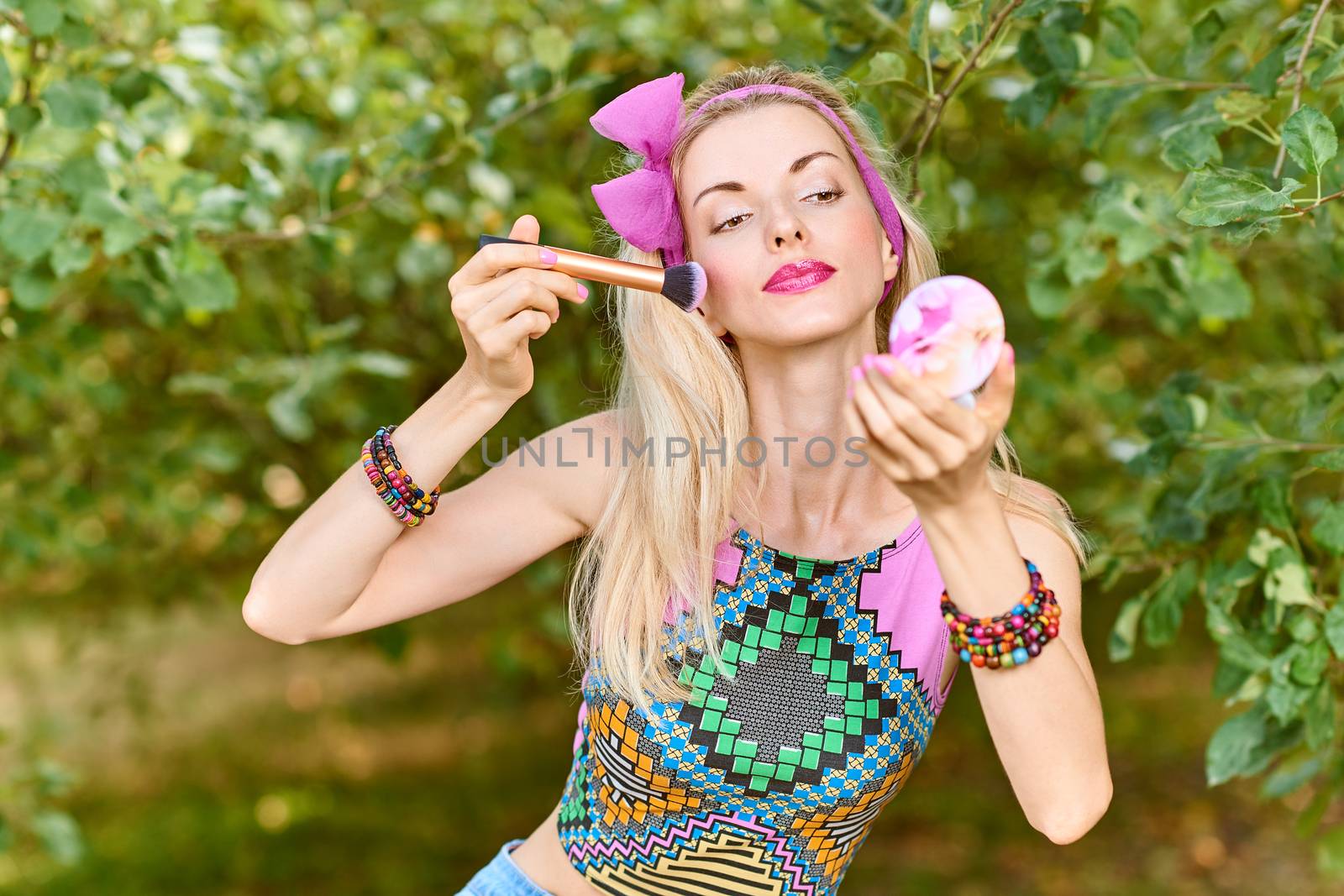 Beauty portrait stylish playful woman smiling primping with mirror, park, people, outdoors. Attractive hipster happy pretty blonde girl with bow, fashionable top. Relax, summer garden, lifestyle,bokeh