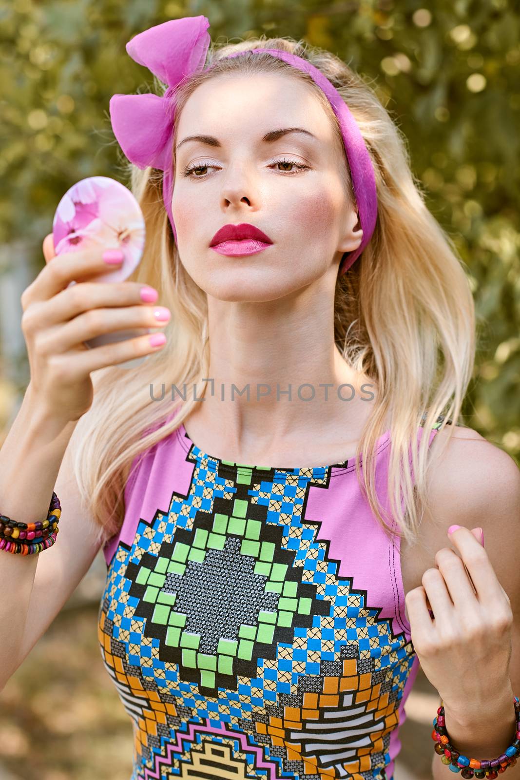 Beauty portrait stylish playful woman primping with mirror in park, people, outdoors. Attractive hipster happy pretty blonde girl with bow, fashionable top. Relax in summer garden, lifestyle, bokeh