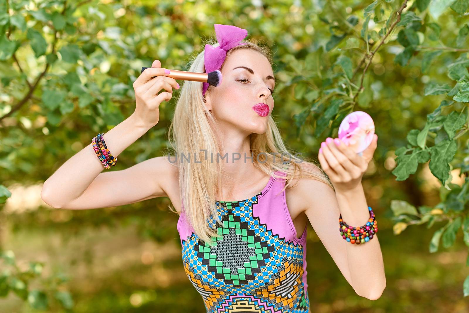 Beauty portrait stylish playful woman primping with mirror in park, people, outdoors. Attractive hipster happy pretty blonde girl with bow, fashionable top. Relax in summer garden, lifestyle, bokeh