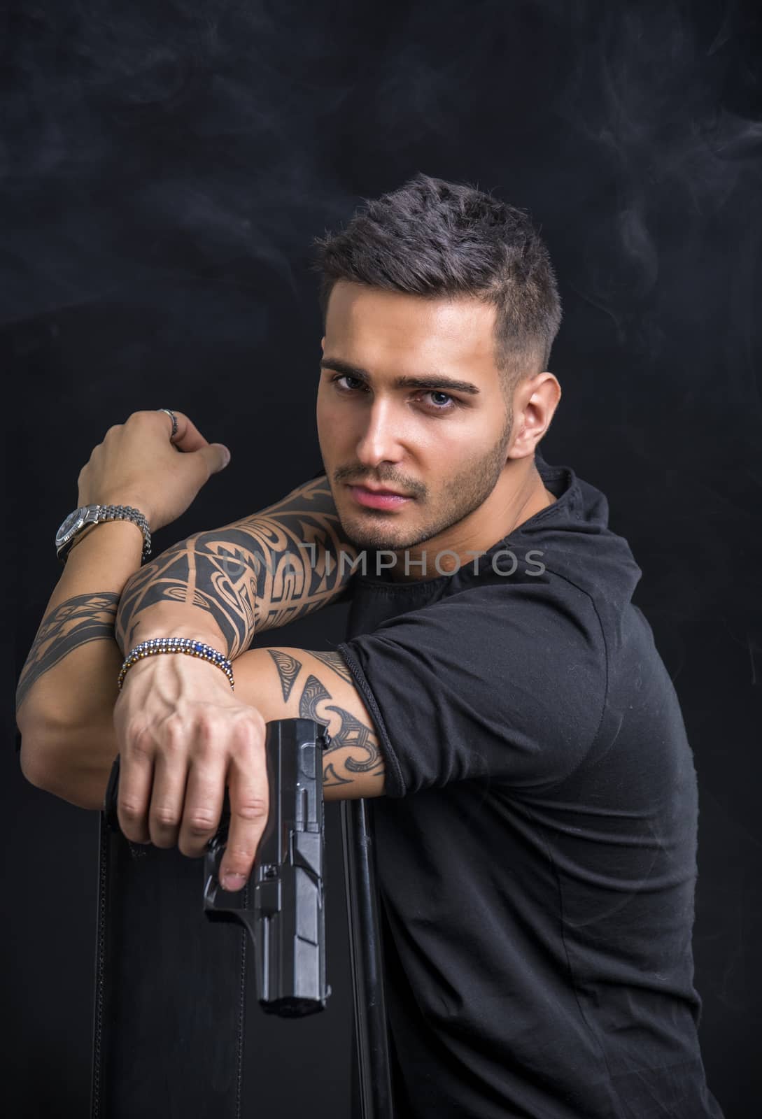 Young handsome man holding a hand gun, wearing black t-shirt, on dark background in studio