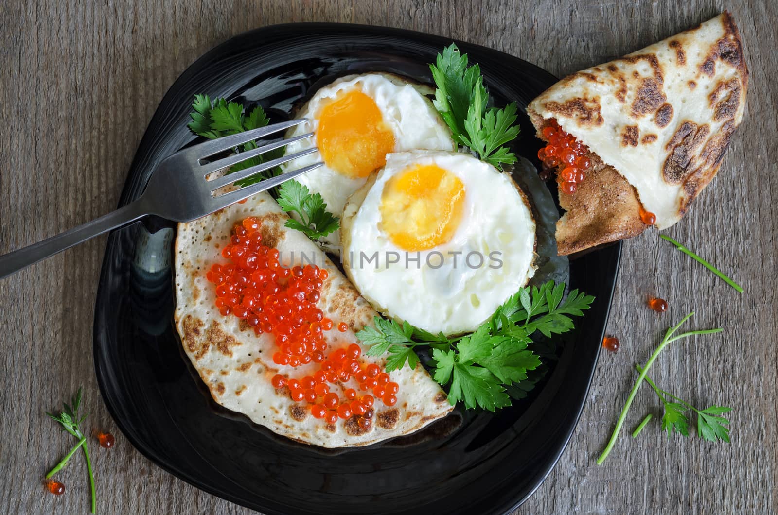Two fried eggs and pancakes with red caviar on a black glass plate, fork and old wooden background