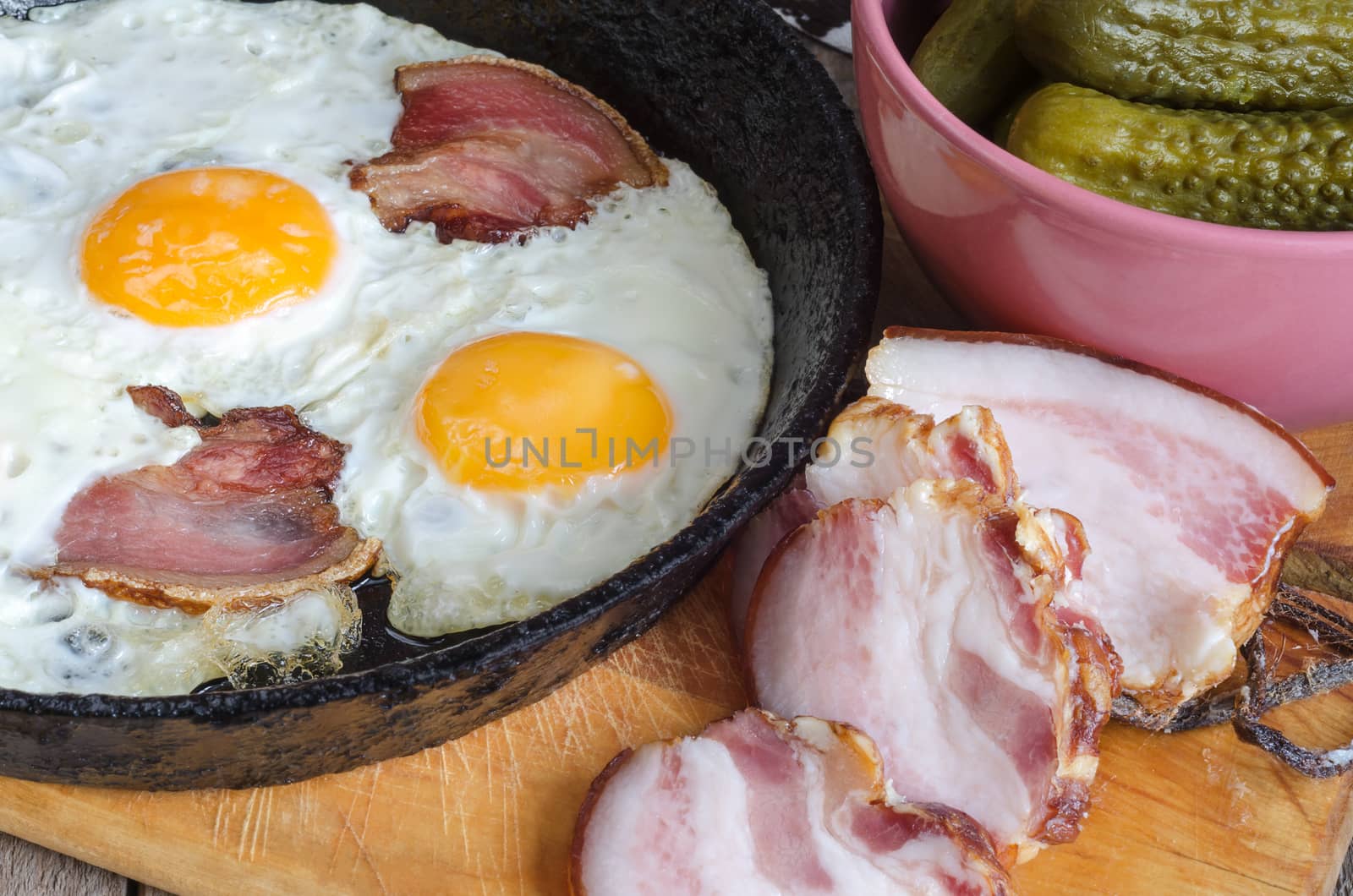 Bacon and eggs in an old pan on the Board. Sliced bacon and pickles in a bowl.
