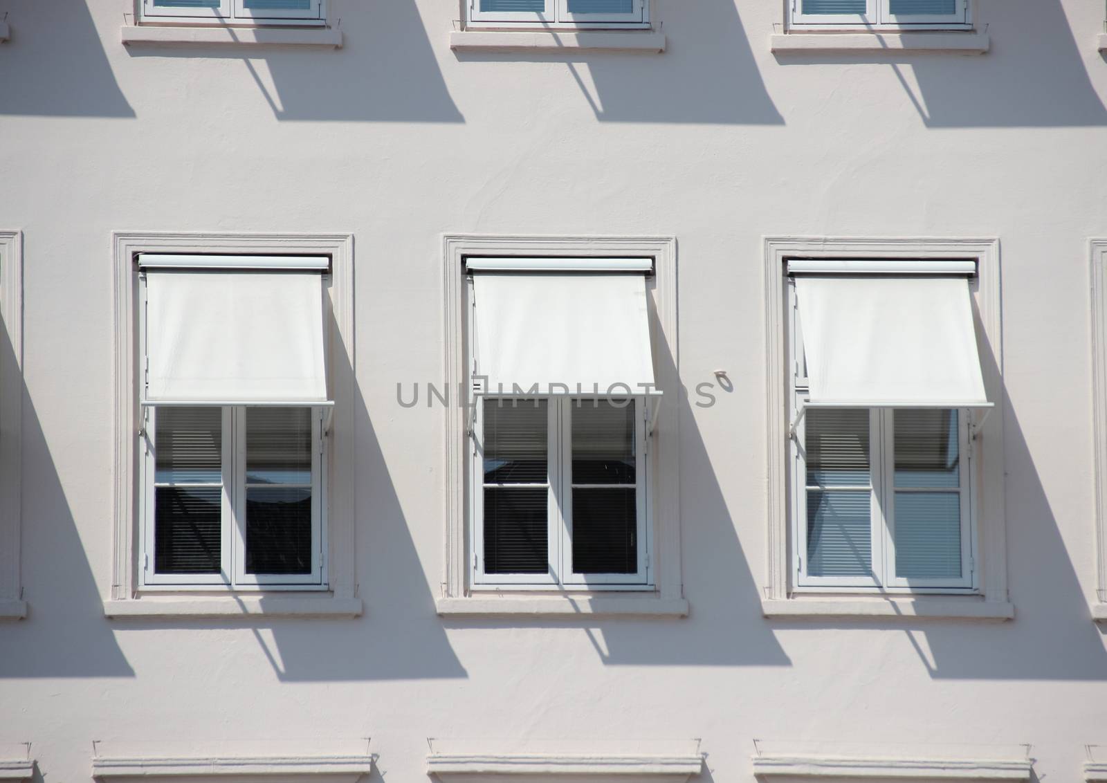 Three windows on grey building with  white awnings and shadow