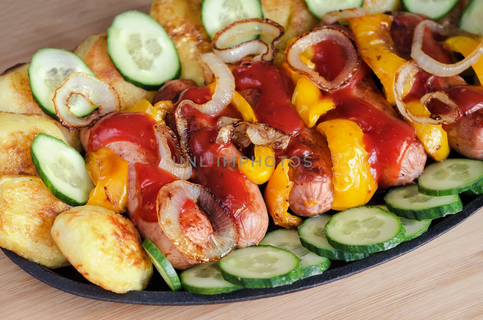 Sausage on a platter with ketchup, roasted potatoes,peppers and onions. Slices of fresh cucumber, wooden background.