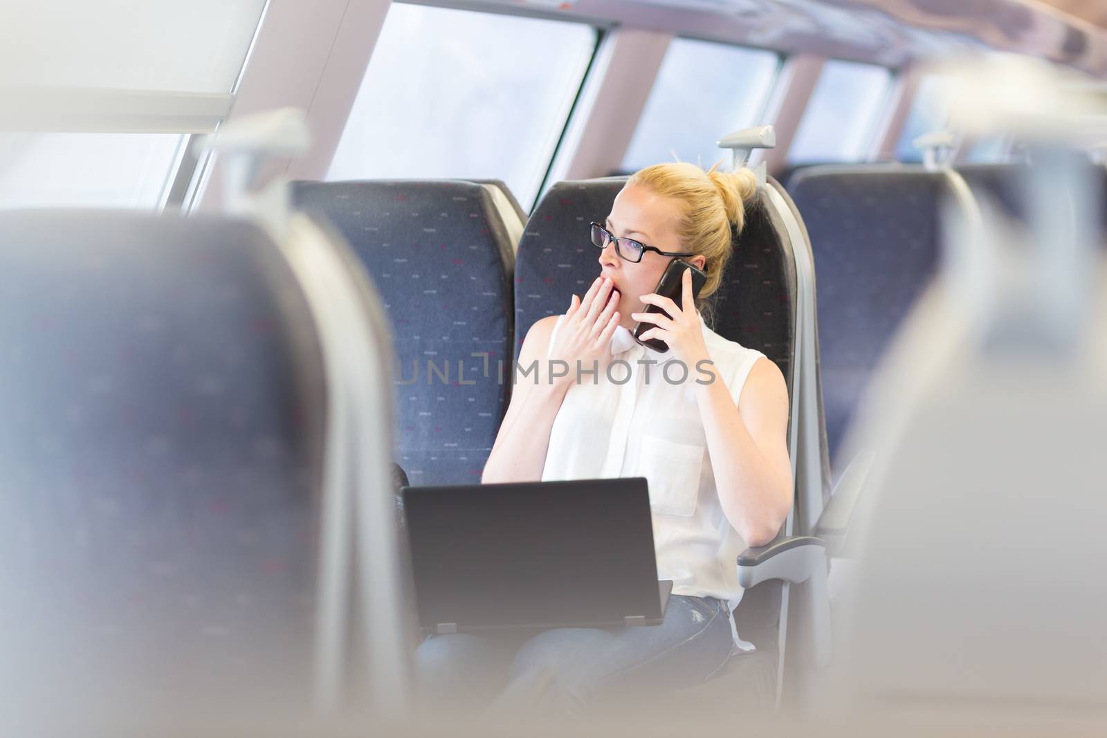 Businesswoman traveling by train, yawning while talking on cellphone and working on laptop. Long and tiresome business travel concept.