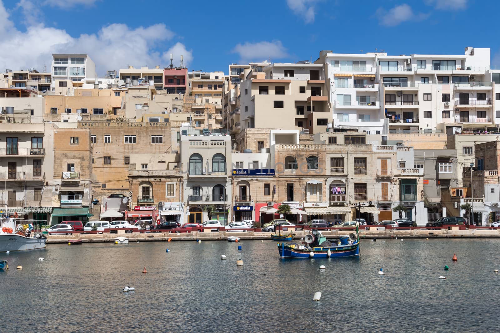 Bay in the city Marsascala (Marsaskala),with a path lining the water of Mediterranean sea in the background. Shops and residential houses. Cloudy sky.