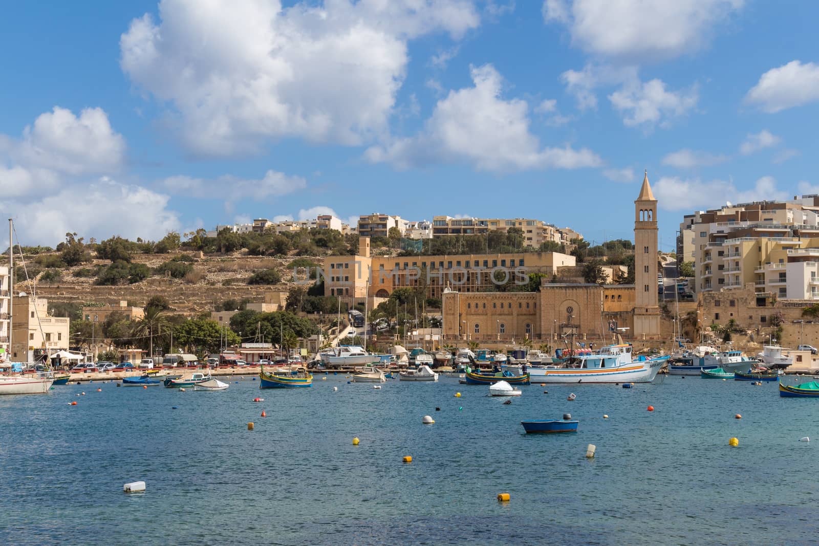 Bay in the city Marsascala,island Malta.Bay of the Mediterranean sea with many fishing boats.Church and houses in the background.Blue sky with white clouds.