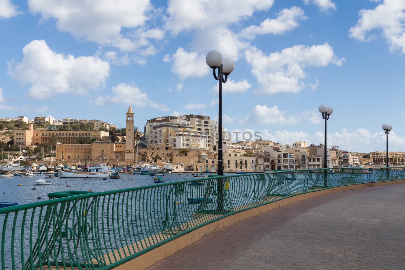 Bay in the city Marsascala (Marsaskala),with a path lining the water of Mediterranean sea. Many fishing boats in the bay. City and a church. Cloudy sky.