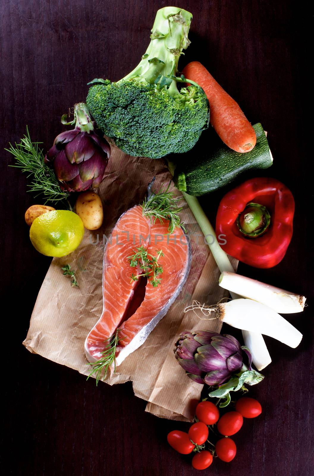 Raw Steak of Salmon on Parchment Paper and Heap of Raw Vegetables with Broccoli, Artichokes, Spring Onion, Tomatoes, Potato, Red Bell Pepper and Lemon closeup on Dark Wooden background. Top View