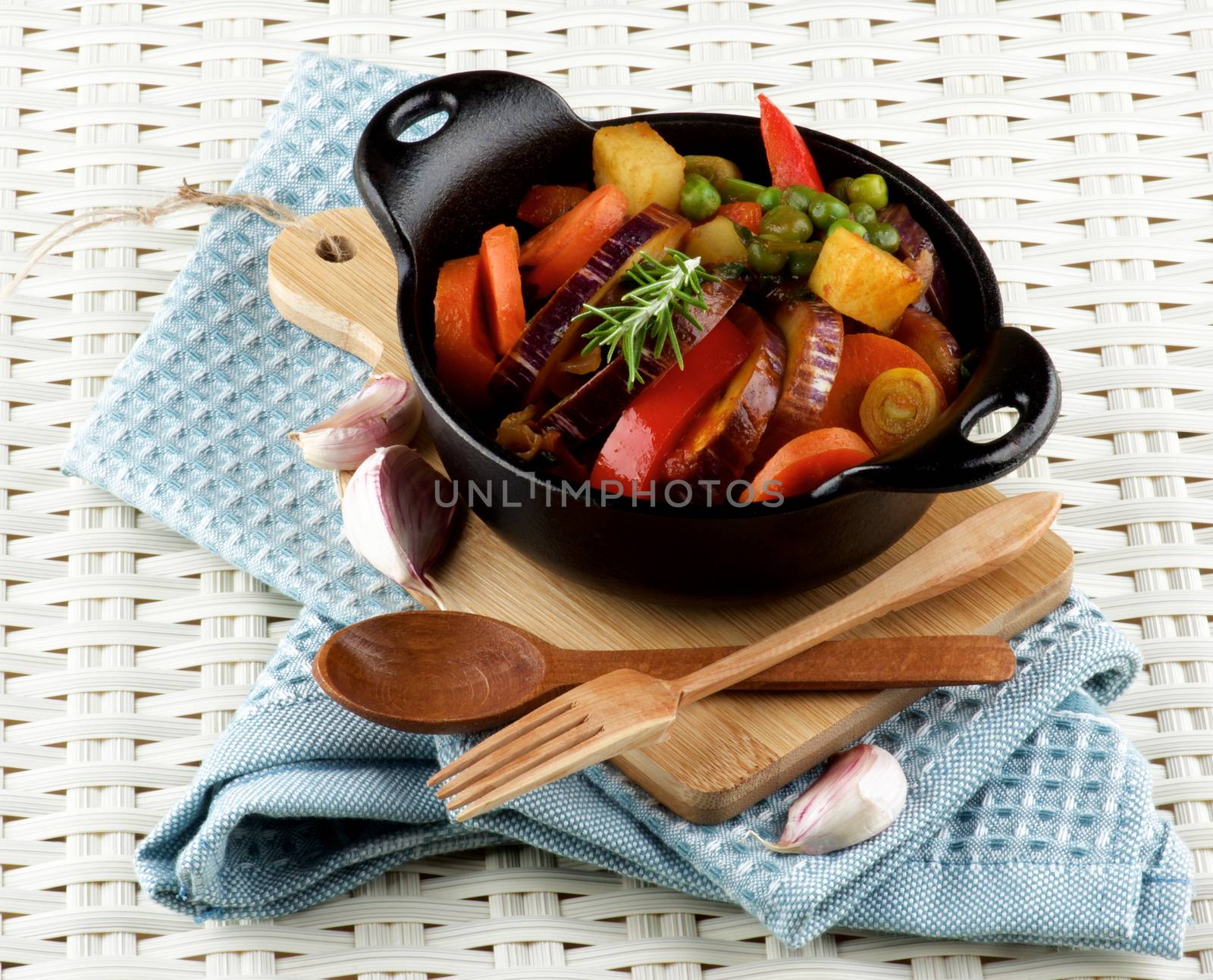 Delicious Homemade Colorful Vegetables Ragout with Striped Eggplant, Carrots, Potatoes, Red Bell Pepper and Green Pea in Black Iron Stewpot with Wooden Fork and Spoon closeup Blue Napkin on Wicker background