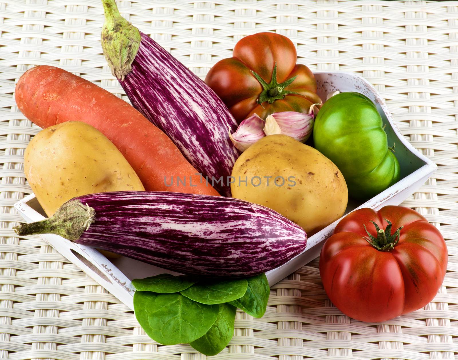 Arrangement of Fresh Raw Vegetables with Striped Eggplants, Potatoes, Green and Red Tomatoes, Carrot, Pink Garlic and Spinach into Wooden Tray closeup on Wicker background