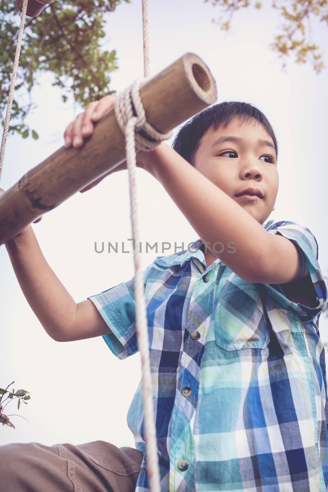 Handsome asian boy climbing on rope ladder made of wood. Child playing outdoors. Travel and adventure concept. Vintage style.