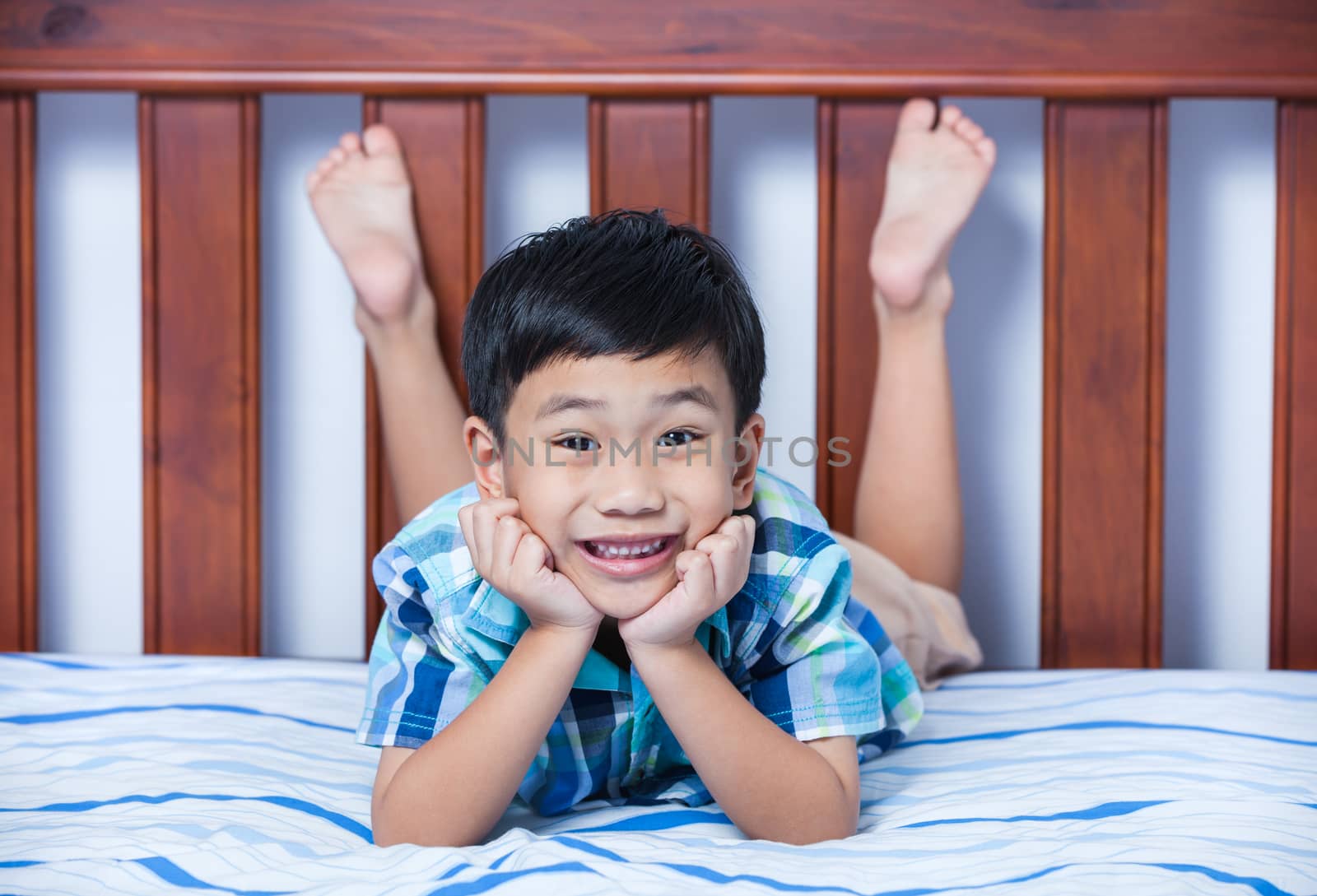 Handsome asian boy with  teeth smile and looking at camera. Lovely child propping up his face and lying barefoot on bed in bedroom.