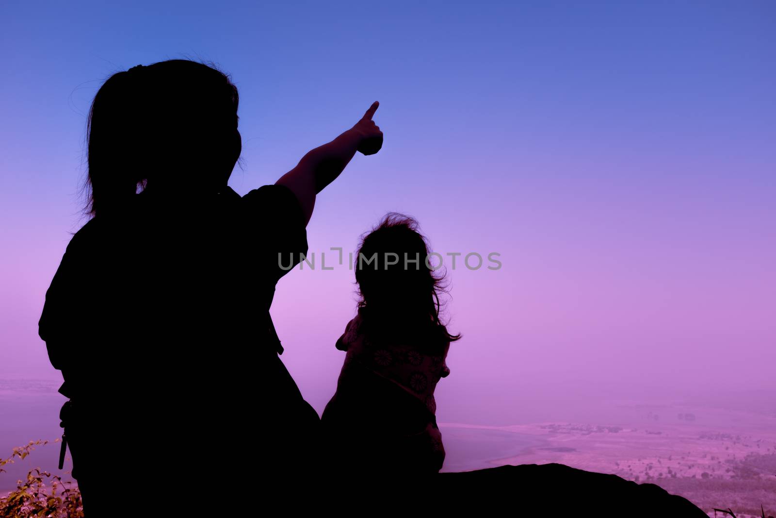 Silhouette back view of young mother and child hikers enjoying the view at the top of a mountain. Woman pointing to empty copy space at sky. Friendly family.