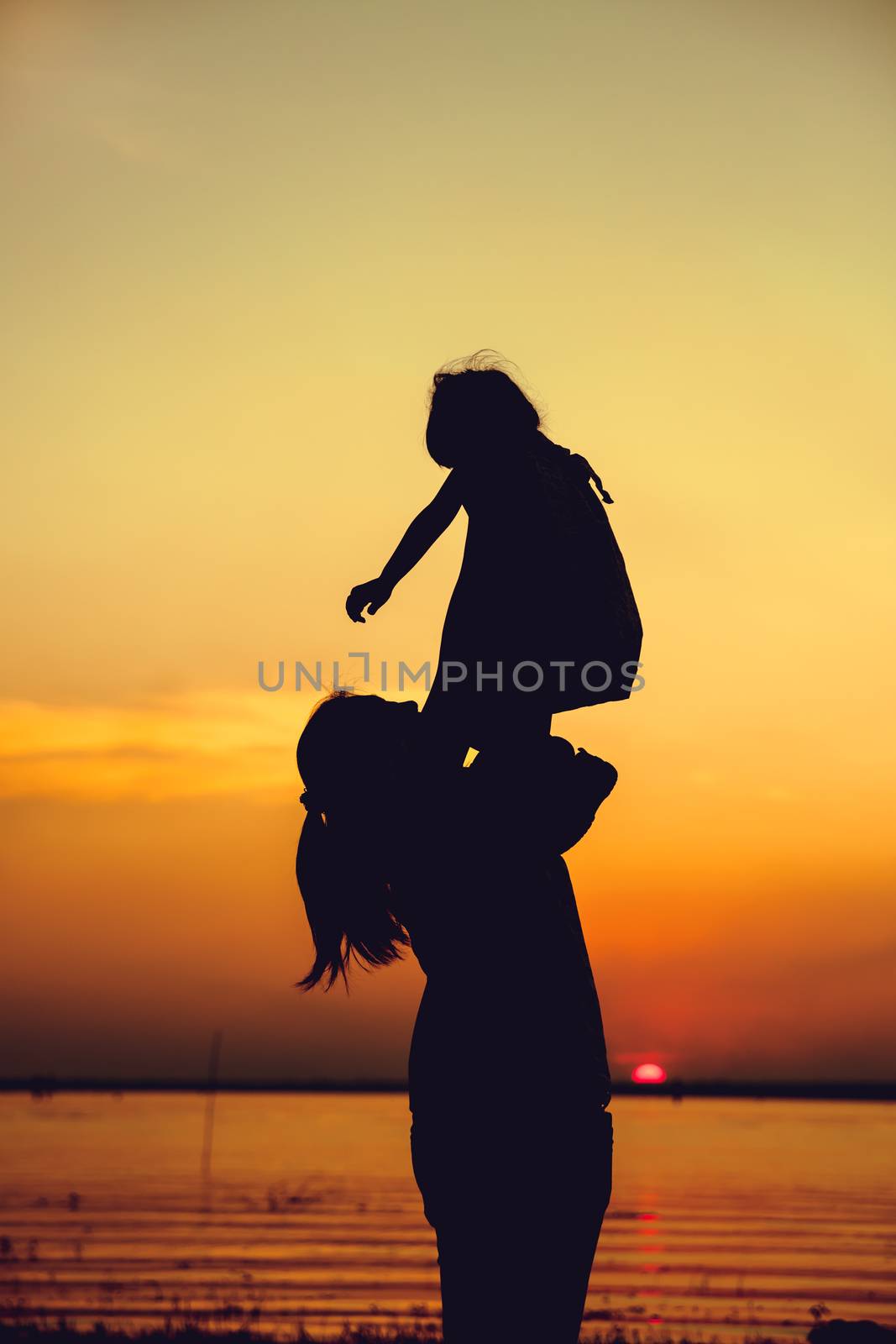 Silhouette of mother and child enjoying the view at riverside. Mother lifting her little girl up in the air on colorful sunset sky background. Friendly family.