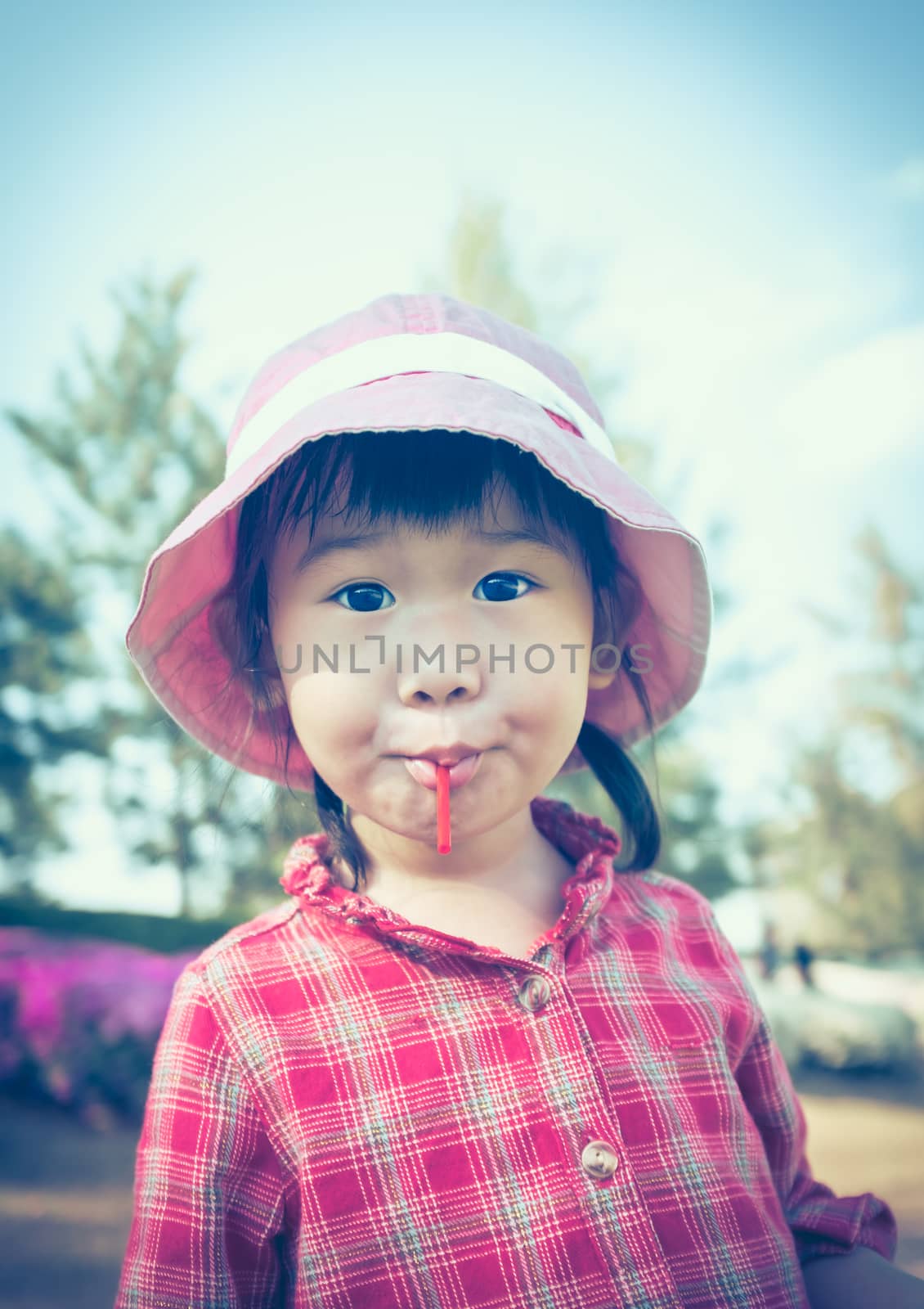 Cute little asian girl eating a lollipop on nature background in summertime. Child wearing pink hat and looking at camera. Cross process. Vintage style.