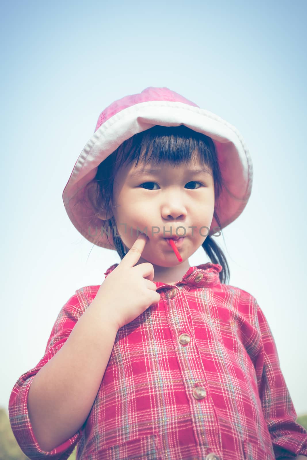 Cute little asian girl eating a lollipop on nature background in summertime. Child wearing pink hat and looking at camera. Cross process. Vintage style.