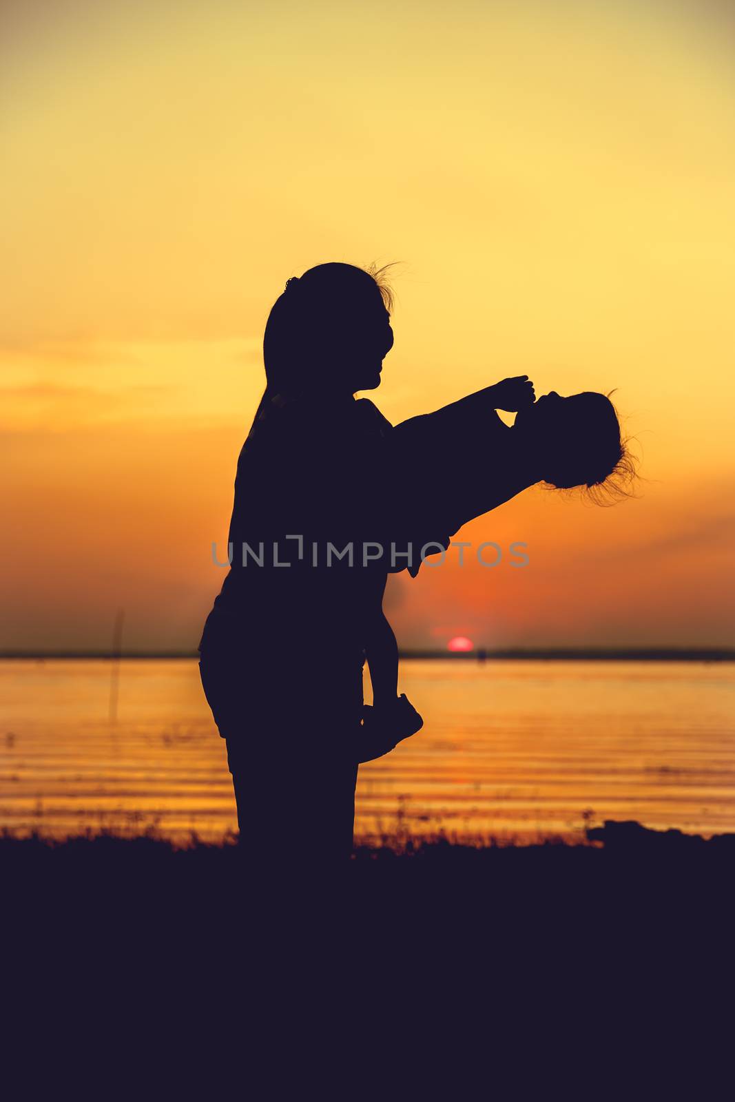 Silhouette of mother and child enjoying the view at riverside. Mother carrying her daughter on colorful sunset sky background. Friendly family.