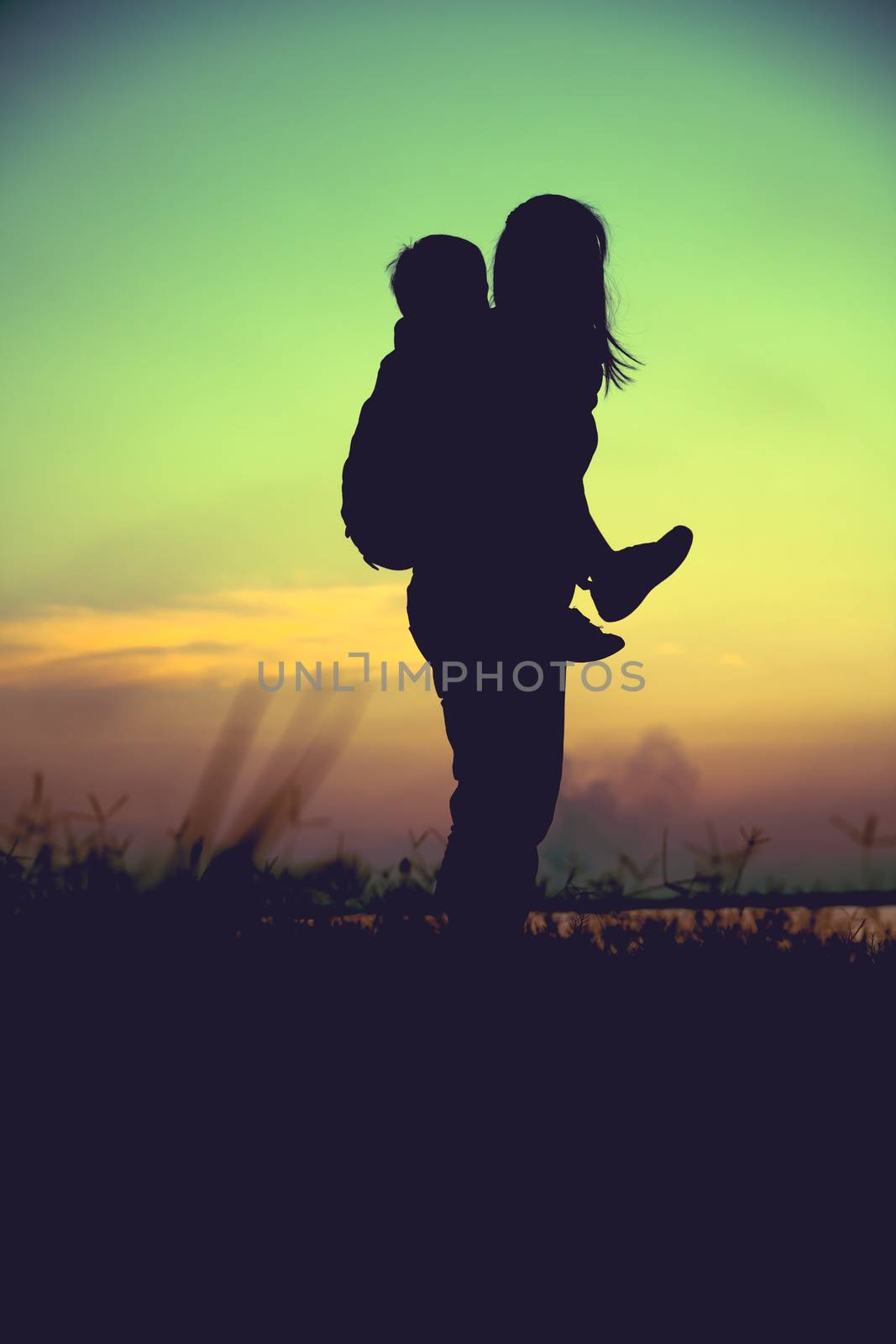 Silhouette of mother and child enjoying the view at riverside. Mother carrying her son on colorful sunset sky background. Friendly family. Cross process. Vignette and vintage style.