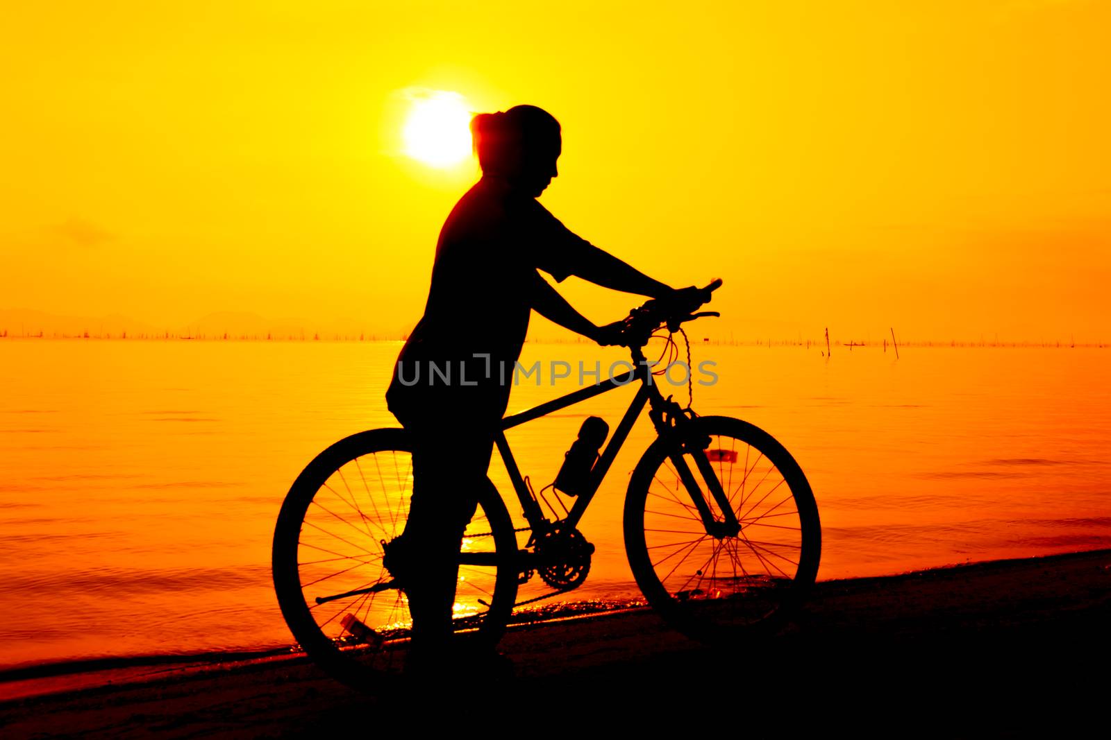 Silhouette of bicyclist enjoying the view at seaside, on colorful sunset orange sky background. Reflection of sun in water. Active outdoors lifestyle for healthy concept.