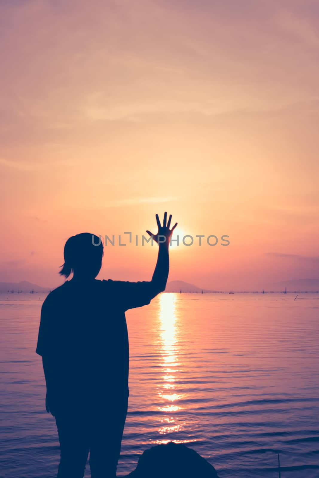 Silhouette back view of woman relax at seaside and showing stop gesture. The sun was hiding from hand on colorful orange sky background. Vintage picture style.