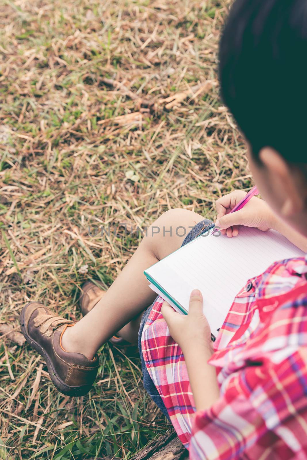 Back view. Child use pen to writing on notebook on wooden log in park. Outdoors in the day time with bright sunlight. Use it for learning or education concept. Vintage style.