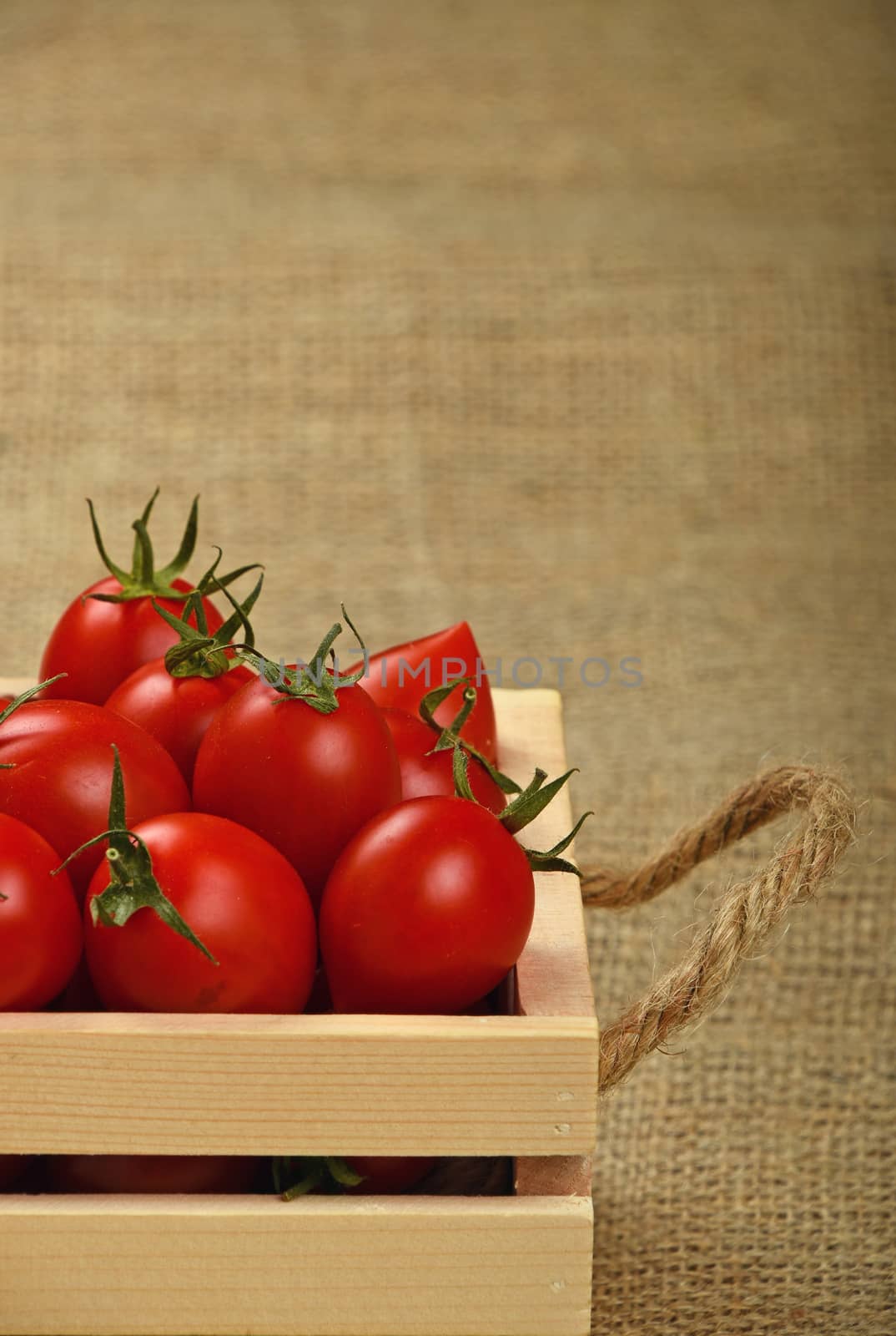 Red ripe fresh cherry tomatoes in small wooden box with twine jute handles on burlap canvas background