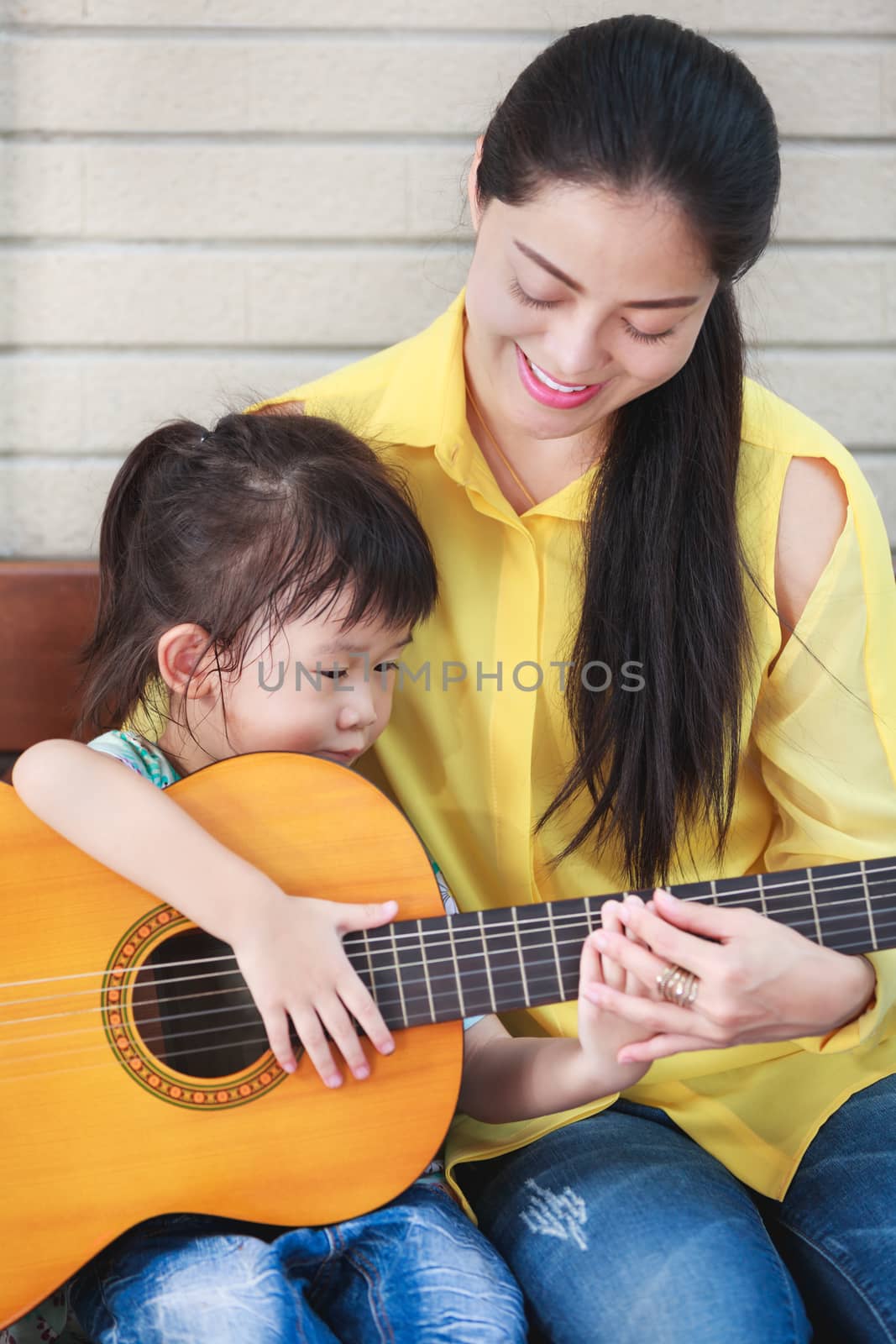 Happy family spending time together at home. Asian mother with daughter playing classic guitar. Positive human emotion.