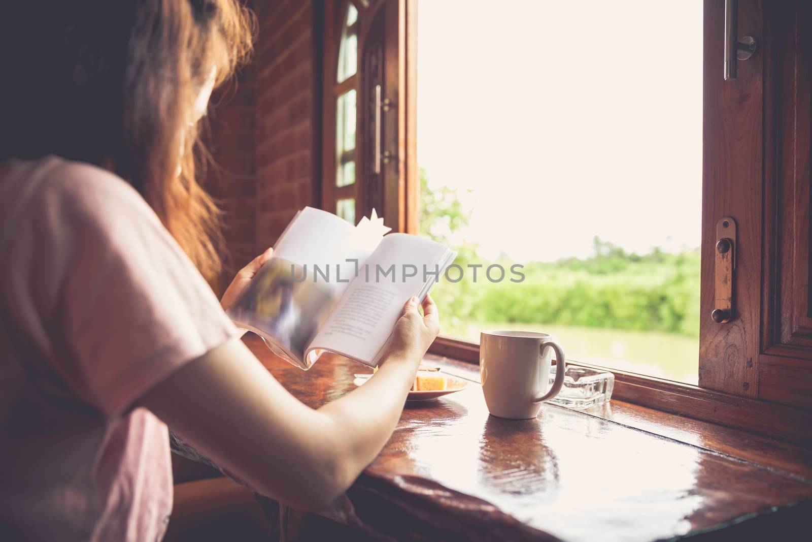 Lifestyle of woman reading a book and enjoy fresh coffee or tea in the morning at home. Sun is shining into the room. Light shines through the window. Happy time of country life. Vintage tone.