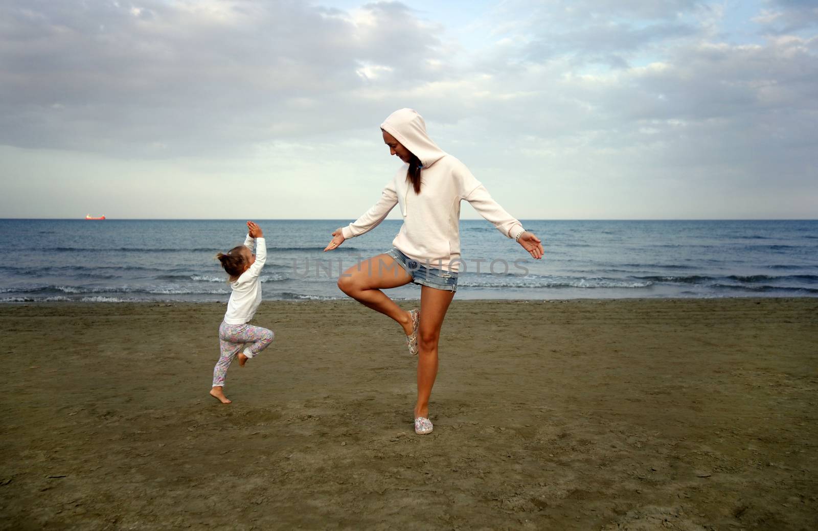 Young woman with small douther practicing yoga on the sea beach at sunset