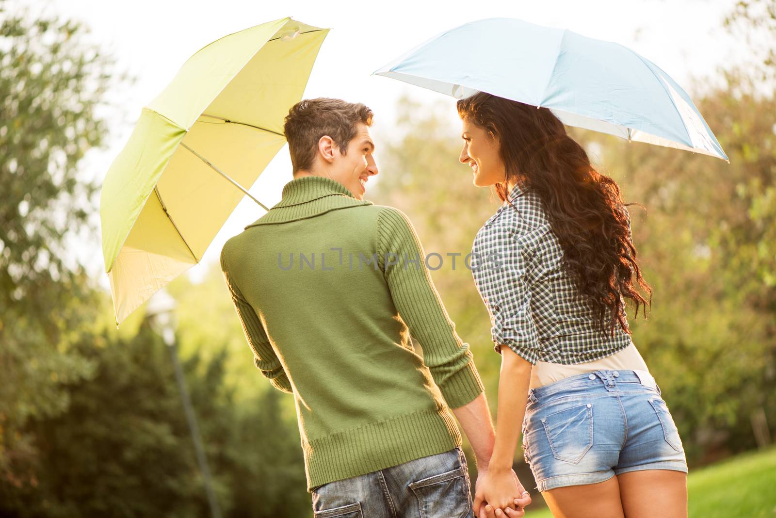 Rear view of a young couple in love with umbrellas while walking through the park holding hands and looking at each other with a smile.