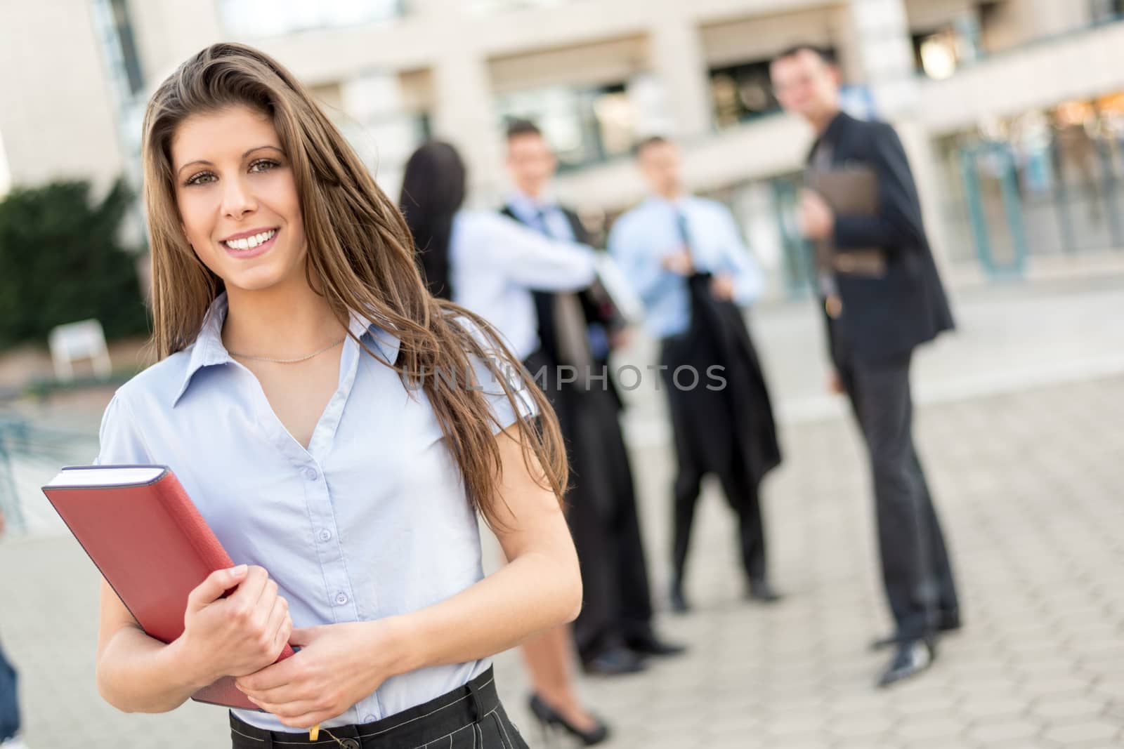 Young businesswoman with planner in her hands, standing in front of office building separated from the rest of the business team. With a smile looking at the camera.